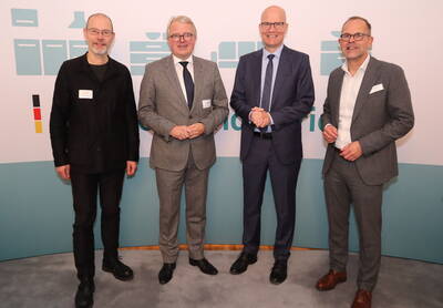 Vier Männer in Anzügen und Namensschildern stehen nebeneinander vor einer hellen Konferenzwand und lächeln in die Kamera