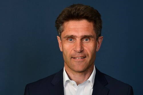 Mittelalterlicher Mann im dunklen Anzug mit weißem Hemd blickt lächelnd in die Kamera vor dunkelblauem Hintergrund