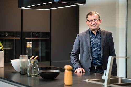 Mittelalterlicher Mann im Anzug lehnt lächelnd an einer modernen Kücheninsel mit Spüle, Kochutensilien und dunklen Einbauöfen im Hintergrund.