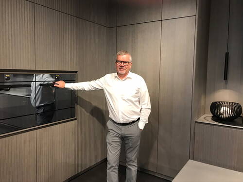 Mann im weißen Hemd demonstriert einen eingebauten Backofen in einer modernen, grauen Einbauküche