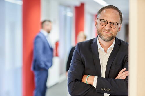 Mittelalterlicher Mann mit Brille und Bart steht mit verschränkten Armen im modernen Büroflur, während unscharf im Hintergrund zwei Personen im Gespräch sind.