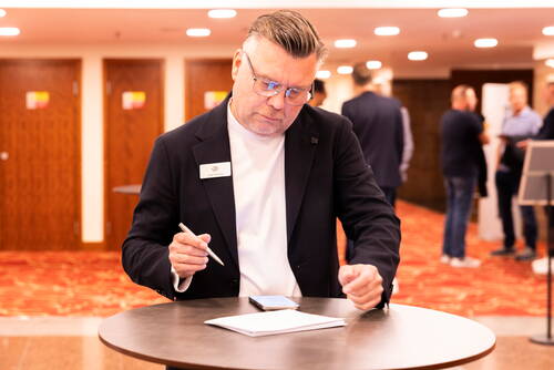 Mittelaltriger Mann mit Namensschild schreibt konzentriert an einem Stehtisch in einer Konferenzlobby, während im Hintergrund mehrere Personen im Gespräch stehen.