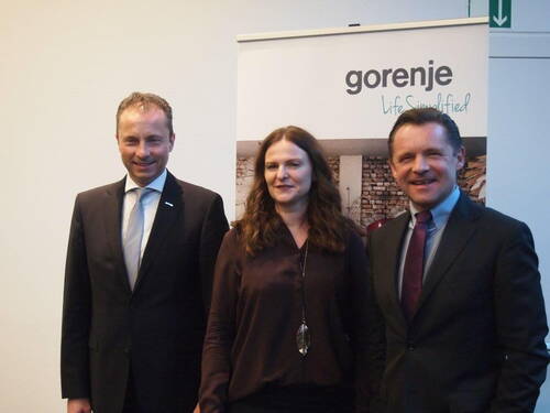 Drei Geschäftsleute posieren lächelnd nebeneinander vor einem Roll-up mit dem Firmenlogo „gorenje“ in einem Konferenzraum.