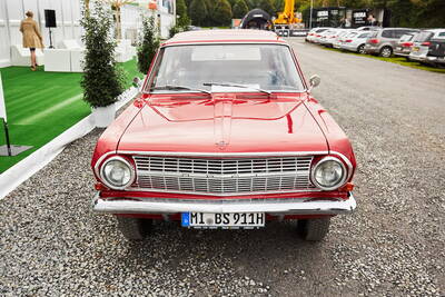 Knallroter, restaurierter Oldtimer mit deutschem Kennzeichen steht auf einem Schotterplatz vor einer Eventfläche mit grüner Kunstrasenfläche und weißen Sitzmöbeln.