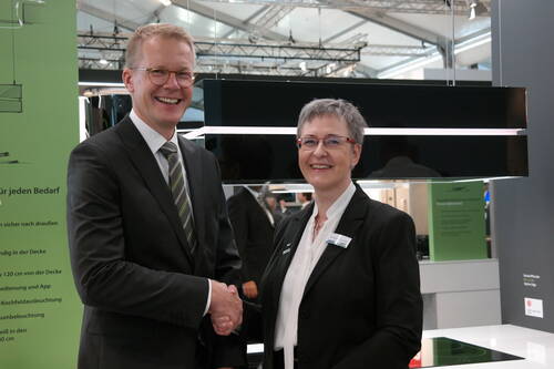 Zwei lächelnde Geschäftsleute in Anzug und Kostüm geben sich auf einer Messe vor einer modernen Küchenausstellung die Hand