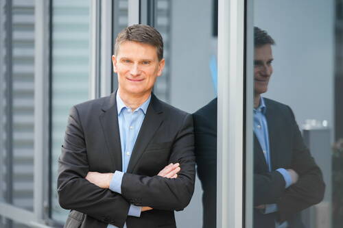 Geschäftsmann im dunklen Anzug steht mit verschränkten Armen lächelnd vor einer Glasfassade, in der sich sein Spiegelbild spiegelt.