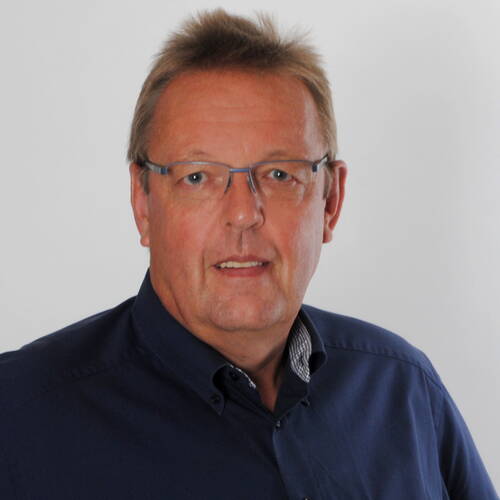 Mittelalterlicher Mann mit Brille und dunkelblauem Hemd blickt freundlich in die Kamera vor neutralem, hellem Hintergrund.