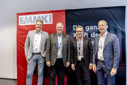 Vier Geschäftsleute im Anzug posieren lächelnd vor einem EMMK-2022-Konferenzbanner in einem Tagungsraum