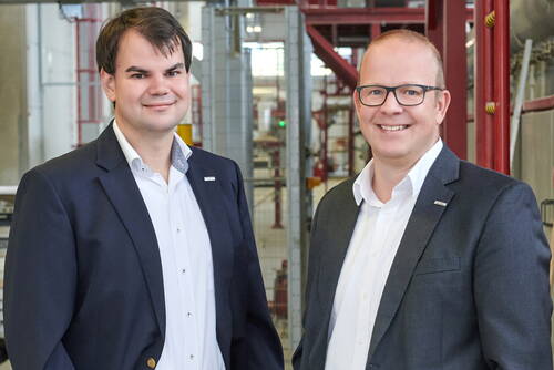 Zwei lächelnde Männer im Business-Anzug stehen nebeneinander in einer modernen Industriehalle mit Maschinen im Hintergrund