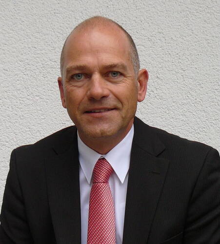 Glatzköpfiger Mann im dunklen Anzug mit weißem Hemd und roter Krawatte lächelt vor einer hellen Putzwand in die Kamera.