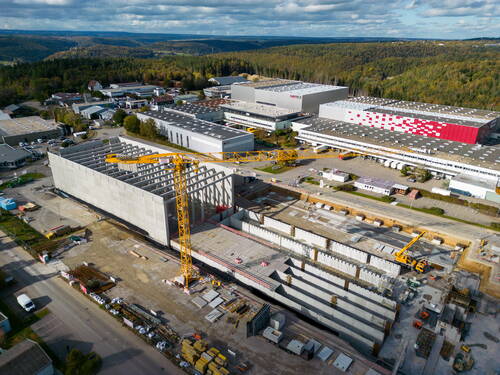 Großbaustelle eines Logistik- oder Industriegebäudes mit gelben Kränen neben bestehenden Hallen am Rand eines bewaldeten Tals
