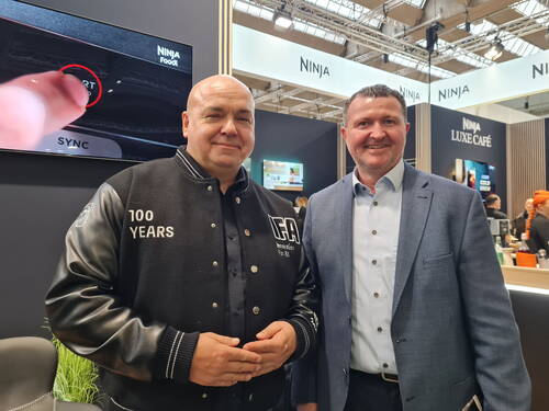 Zwei lächelnde Männer posieren auf einer Messe vor einem Ninja-Foodi-Stand, einer trägt eine schwarze Collegejacke mit der Aufschrift „100 Years“, der andere einen blauen Anzug.