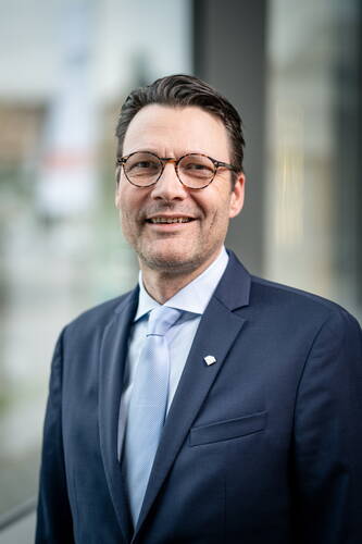 Lächelnder Mann mit Brille im dunkelblauen Anzug und hellblauer Krawatte vor unscharfem, modernem Hintergrund