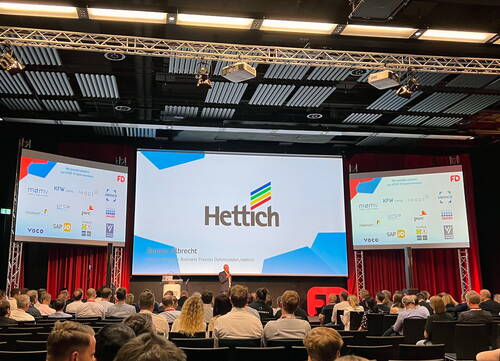 Geschäftspräsentation von Hettich vor großem Publikum in einem Konferenzsaal mit drei Projektionsleinwänden und einem Redner auf der Bühne