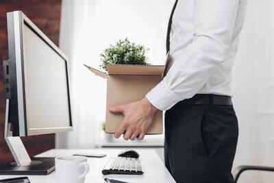 Angestellter im Business-Outfit trägt einen Karton mit Schreibtischpflanze und räumt seinen Arbeitsplatz vor einem Computer leer