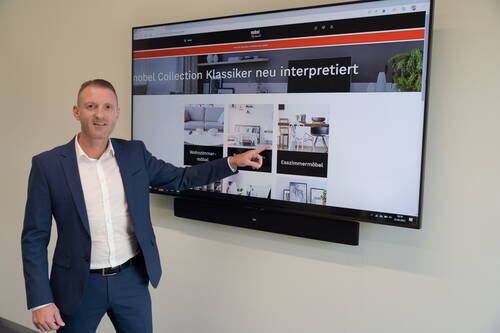 Geschäftsmann im blauen Anzug präsentiert an einem großen Wandbildschirm eine Website mit verschiedenen Möbelkategorien