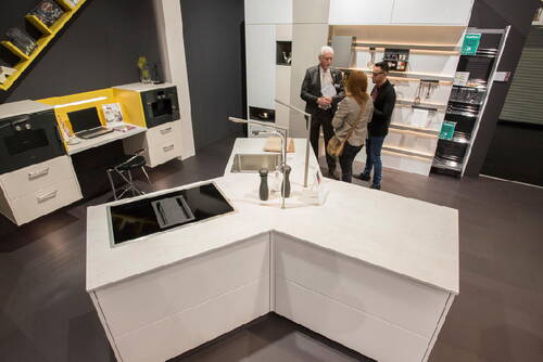 Drei Personen unterhalten sich in einer modernen Ausstellungsküche mit weißer Kochinsel, integriertem Induktionskochfeld, Spüle und angrenzendem Arbeitsplatzbereich.