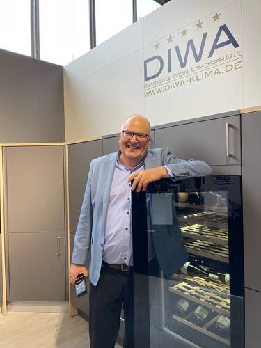Lächelnder Mann im blauen Sakko lehnt sich an einen beleuchteten Weinkühlschrank in einem modernen Ausstellungsraum der Marke DIWA