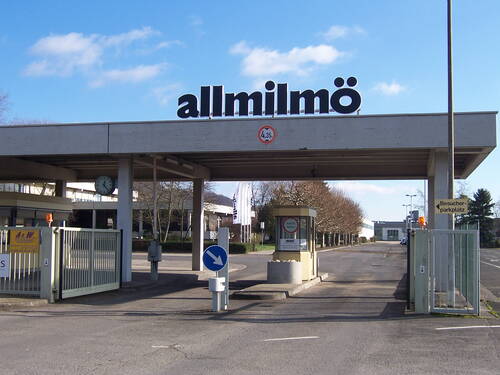 Werkszufahrt mit Pförtnerhäuschen und Schranke unter einem großen Vordach mit der Aufschrift „allmilmö“, flankiert von Zufahrtsstraßen und Besucherparkplätzen an einem klaren Tag.