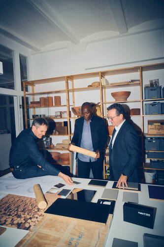 Drei Designer begutachten gemeinsam Materialmuster und Holzproben auf einem großen Arbeitstisch in einem Studio mit Regalen voller Schalen, Modelle und Kisten im Hintergrund.