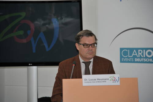 Mann im braunen Sakko spricht nachdenklich am Rednerpult mit Mikrofon vor Präsentationsbildschirm und Veranstaltungslogo