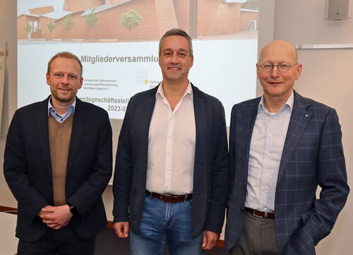 Drei Anzugträger stehen lächelnd vor einer Präsentationsfolie mit der Aufschrift „Mitgliederversammlung“ in einem Konferenzraum