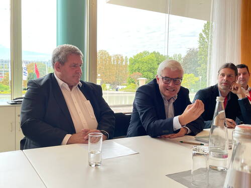 Mehrere Geschäftsleute sitzen an einem Konferenztisch vor einer großen Fensterfront, während der Mann in der Mitte gestikulierend spricht und die anderen aufmerksam zuhören.