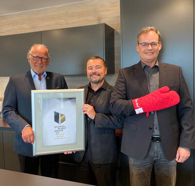 Drei lächelnde Männer in Anzügen präsentieren in einer modernen Küche eine gerahmte German Brand Award Urkunde, während der rechte Mann demonstrativ einen roten Ofenhandschuh trägt