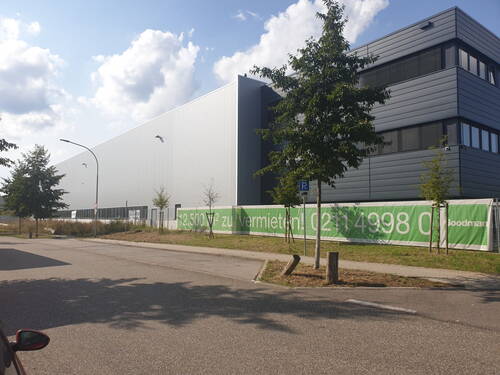 Große moderne Industrie- und Lagerhalle an einer ruhigen Straße mit grünem Banner „12.500 m² zu vermieten“ entlang des Zauns und vereinzelten jungen Bäumen im Vordergrund