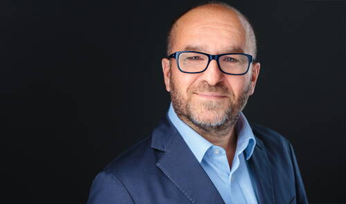 Glatzköpfiger Mann mit Bart und dunkler Brille lächelt leicht in einem professionellen Porträt vor dunkelgrauem Hintergrund und trägt ein blaues Sakko mit hellblauem Hemd.