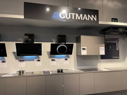 Moderne Küchenausstellung mit GUTMANN-Dunstabzugshauben in Schwarz, beleuchtetem Runddesign und grauen Unterschränken vor hellgrauer Wand mit Informationsblättern