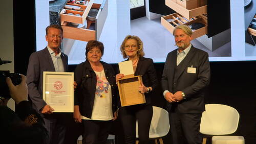 Vier Personen stehen auf einer Bühne und präsentieren einen Interior Innovation Award 2025 sowie ein hölzernes Schubladenmodell vor einer großen Leinwand mit Küchenstauraum-Visualisierungen.