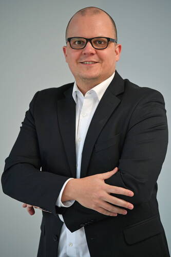 Mann mit schwarzer Brille im dunklen Anzug steht vor neutralem Hintergrund und verschränkt lächelnd die Arme.