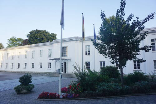 Weißes zweistöckiges Verwaltungsgebäude mit Flaggenmasten und bepflanztem Vorplatz an einem klaren Sommertag