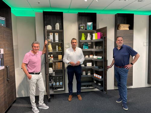 Drei Männer präsentieren in einem modernen Showroom zwei geöffnete Vorratsschränke mit ordentlich arrangierten Küchenutensilien und Lebensmitteln.