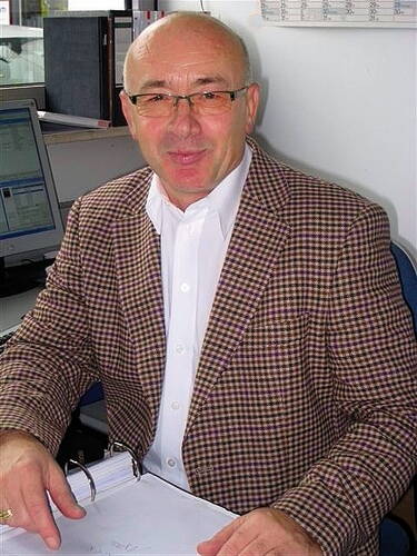 Älterer Mann mit Brille im karierten Sakko sitzt am Schreibtisch und arbeitet an einem geöffneten Ordner vor dem Computer