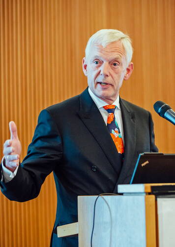 Älterer Mann im Anzug mit bunter Krawatte spricht gestikulierend an einem Rednerpult mit Mikrofon und Laptop in einem Konferenzraum.