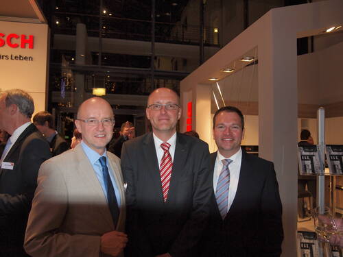 Drei lächelnde Geschäftsmänner in Anzügen posieren auf einer Abendveranstaltung in modern beleuchteten Messe- oder Firmenräumen mit Bosch-Logo im Hintergrund.