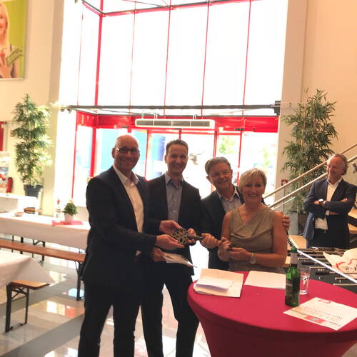 Vier elegant gekleidete Personen lächeln und halten gemeinsam einen symbolischen Schlüssel bei einer feierlichen Übergabe in einem lichtdurchfluteten Foyer mit roten Fensterrahmen.