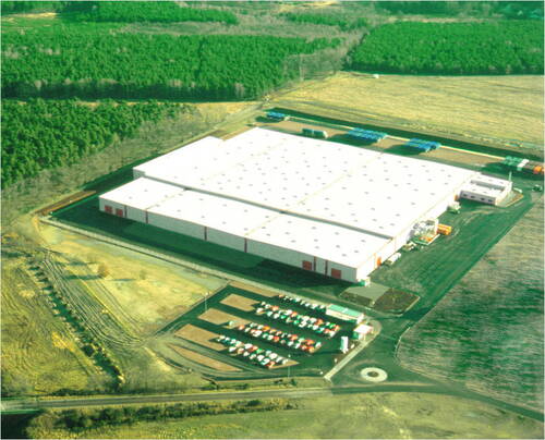 Großes, weißes Logistik- und Lagergebäude mit angrenzendem Parkplatz und Lkw-Flächen, umgeben von Feldern und Wald aus der Luftperspektive.