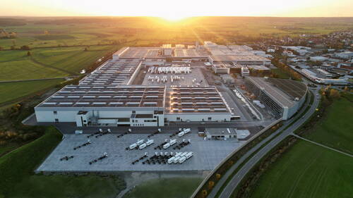 Großes Industrie- und Logistikzentrum mit Solaranlagen auf den Hallendächern, Lkw-Aufliegern auf dem Vorplatz und umliegender grüner Landschaft im Abendlicht