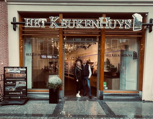 Zwei lächelnde Personen stehen vor dem Eingang des Küchenstudios „Het KeukenHuys“ in einer nassen Einkaufsstraße.