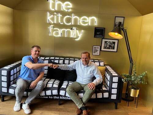 Zwei Männer sitzen einander zugewandt auf einem gemusterten Sofa in modernem Loungebereich und begrüßen sich mit einem Fauststoß vor einem leuchtenden „the kitchen family“-Schriftzug an der Wand.