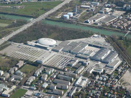 Luftaufnahme eines weitläufigen Messe- und Veranstaltungsgeländes mit großer Kuppelhalle, umliegenden Parkplätzen, Wohngebäuden im Vordergrund und einem Fluss mit Industrieanlagen im Hintergrund