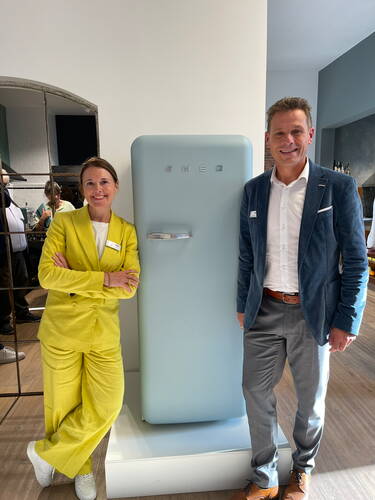 Zwei lächelnde Geschäftsleute stehen in moderner Ausstellung neben einem pastellblauen SMEG-Kühlschrank, sie in gelbem Hosenanzug mit verschränkten Armen, er in blauem Sakko und grauer Hose.