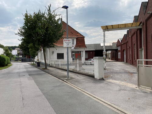 Schmale Industrie­straße mit weißem Bürogebäude und rotem Ziegelhallenkomplex, Einfahrt mit weißem Zaun und OFA-Beschilderung unter bewölktem Himmel.