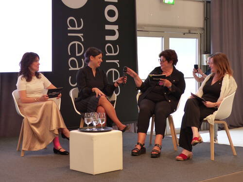 Vier Frauen sitzen auf einer Bühne in einer Podiumsdiskussion, tauschen sich mit Mikrofonen und Notizkarten aus, während im Hintergrund jemand mit dem Smartphone fotografiert.