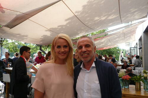 Blonde Frau und älterer Mann lächelnd im Vordergrund einer sonnigen Gartenveranstaltung mit vielen Gästen unter großen Sonnensegeln