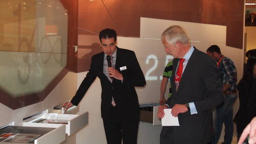 Messemitarbeiter im Anzug erklärt einem älteren Besucher mit roter Krawatte die Funktionsweise ausziehbarer Schubladensysteme an einem modernen Ausstellungsstand.