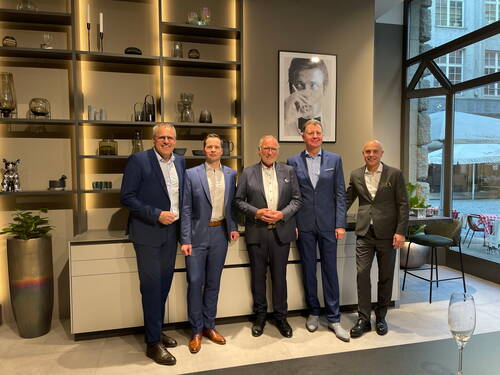Fünf elegant gekleidete Männer posieren lächelnd in einem modern eingerichteten Showroom mit beleuchteten Regalen und großem Fenster zur Straßenansicht.